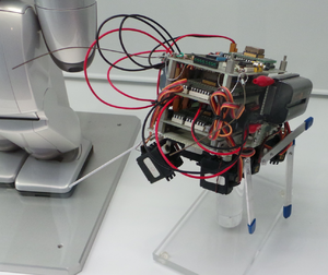 A photograph of the MUTANT prototype mounted on a stand. It is a small, cube-shaped robot on stilts with a lot of protruding red and orange wires.