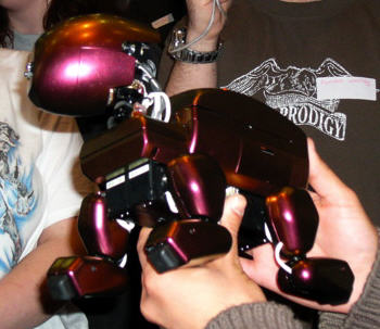 A photograph of the red robodog prototype being held in a tan-skinned person's hands. It is quadrapedal with chunky, rounded body parts and a particularly oblong head.